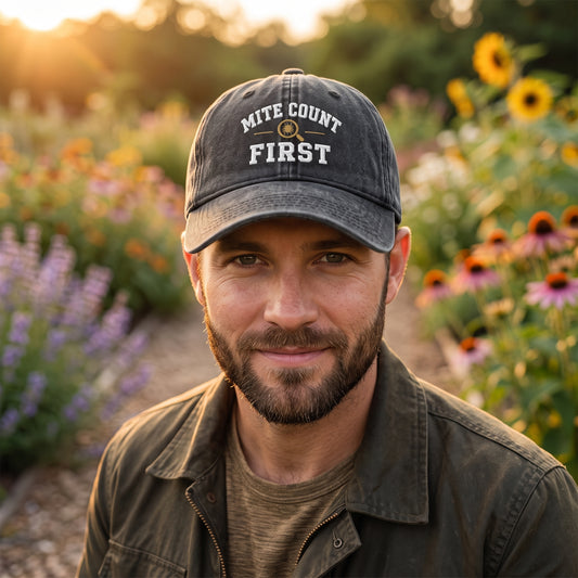 A man wearing a dark gray baseball cap with 'MITE COUNT FIRST' embroidered on the front, standing in a sunlit garden with flowers in the background.
